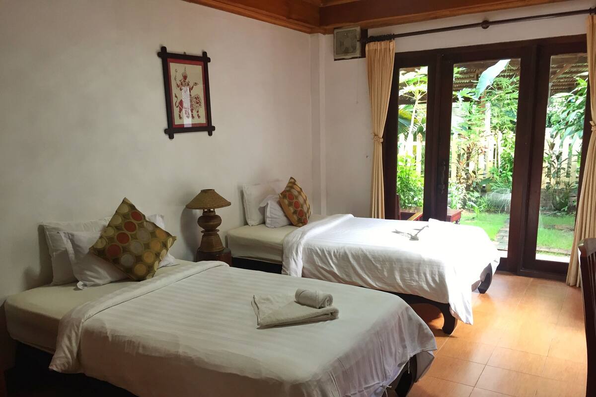 A room with two beds, a framed picture on the wall, and a view of greenery outside.