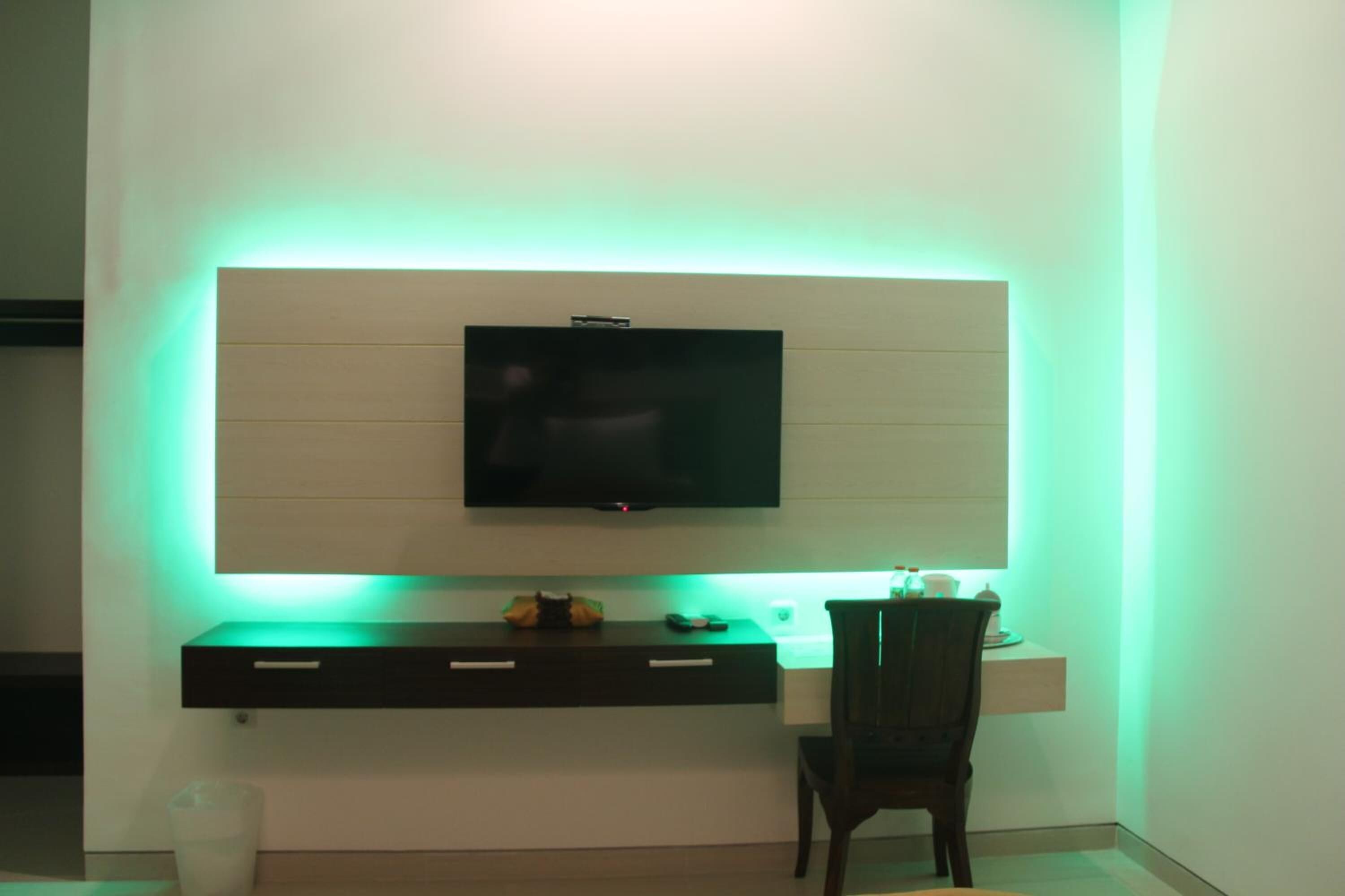 Una habitación moderna con una televisión de pantalla plana montada en una pared blanca, un escritorio de madera oscura con cajones y una sola silla.