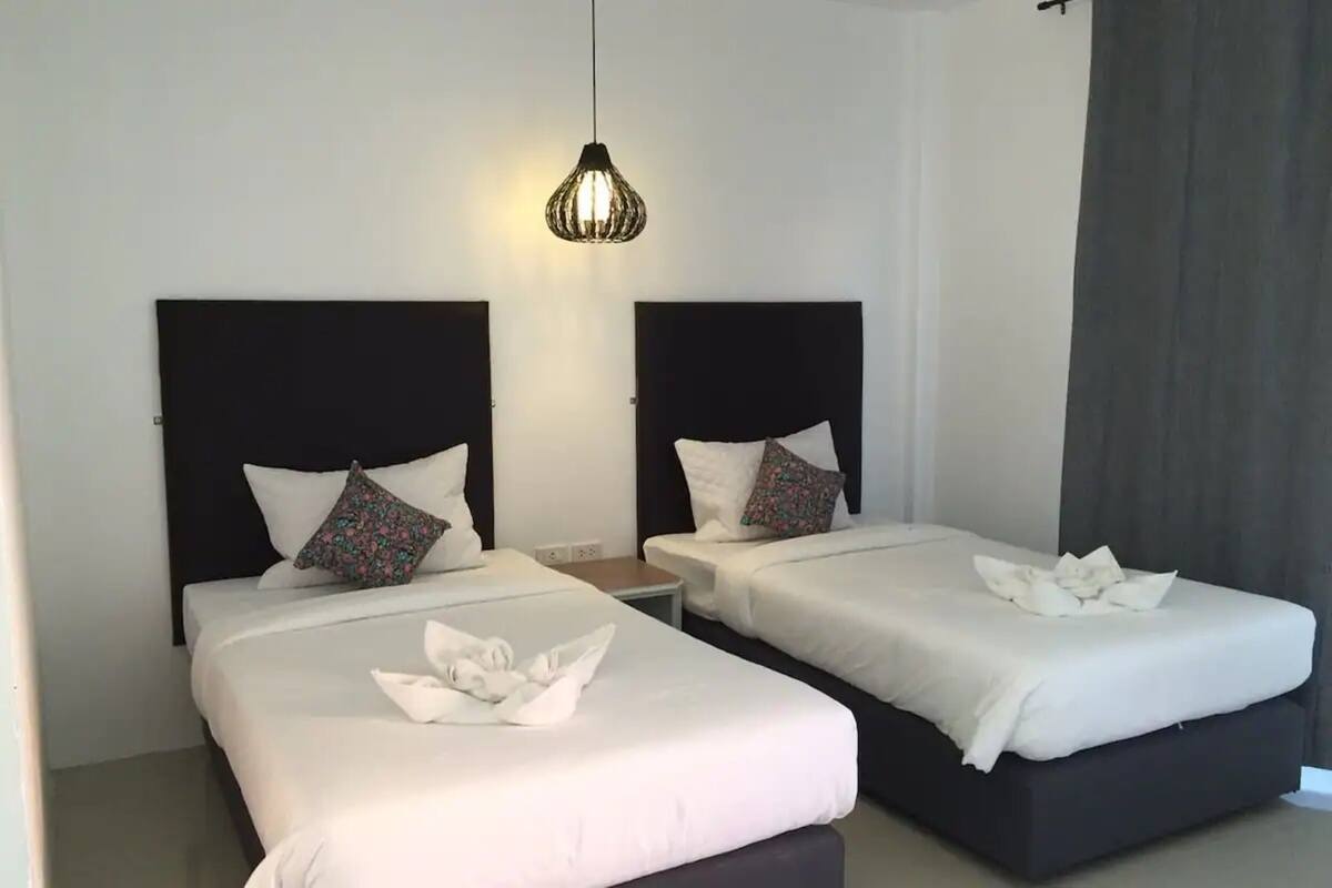 A hotel room with two beds, each with a black headboard and white bedding. There is a small bedside table between the beds, and a single hanging light fixture above.