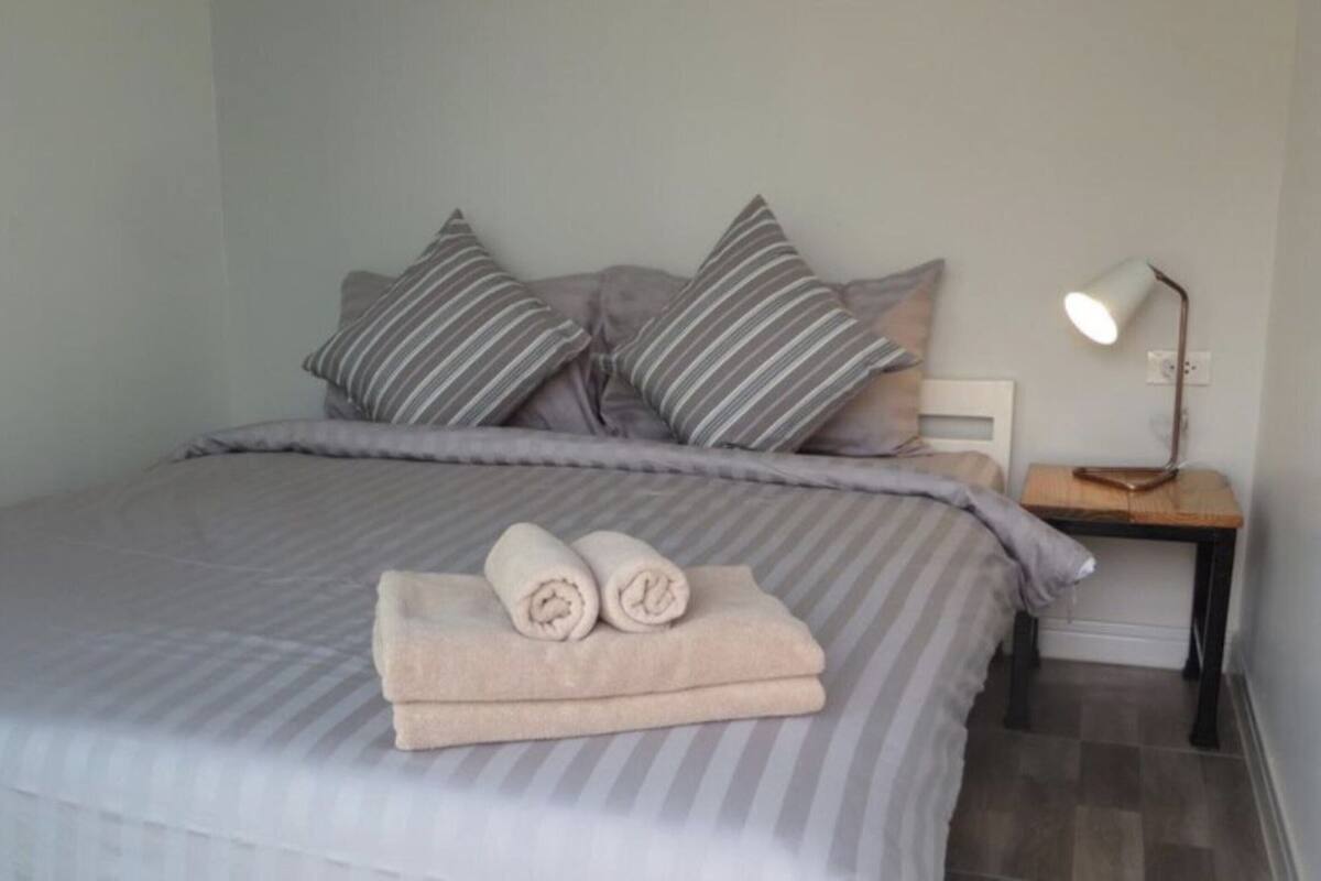 A neatly made bed with striped bedding and pillows, a bedside table with a lamp, and rolled towels on the bed.