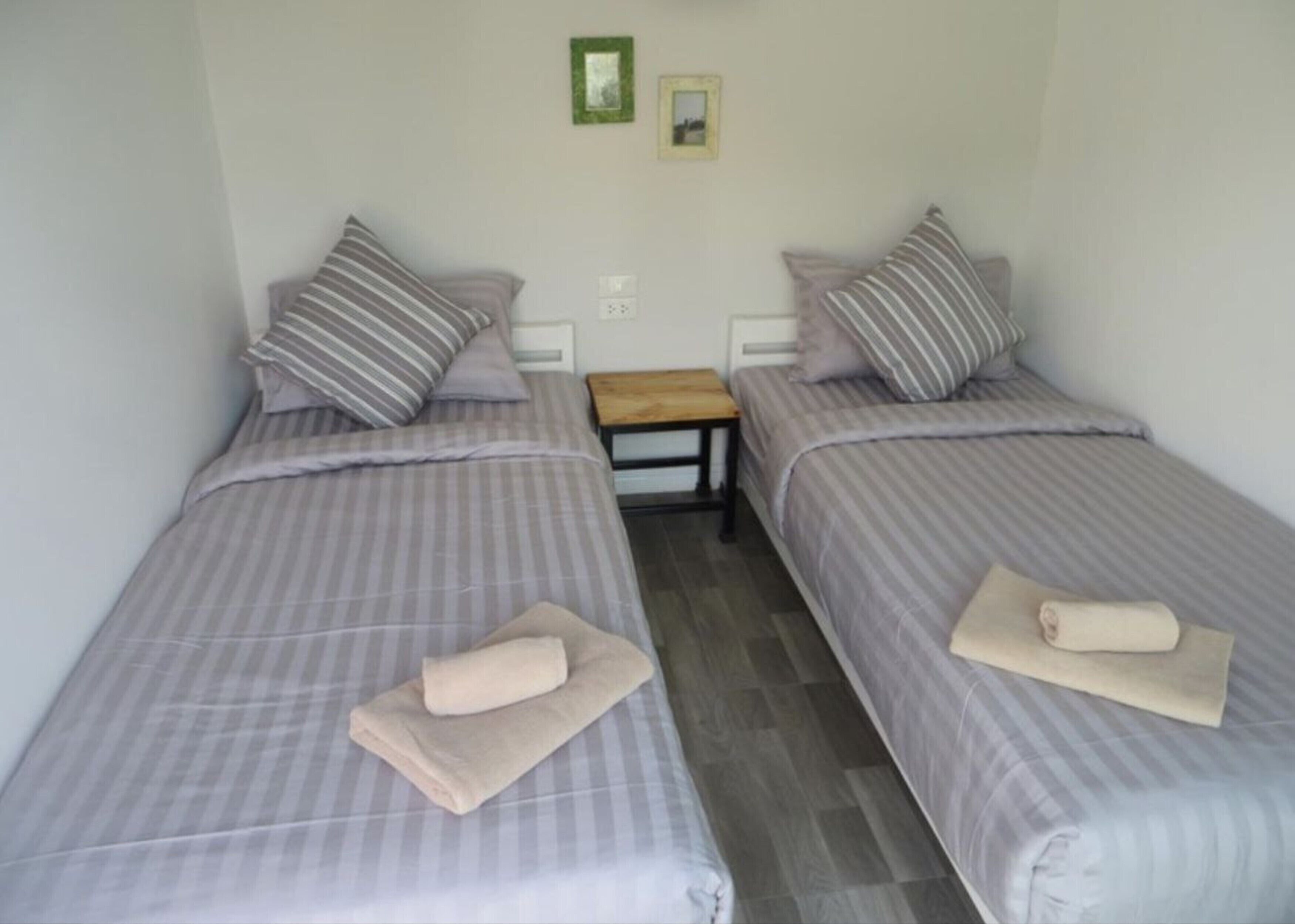 A room with two single beds, each with striped bedding and pillows, a small wooden nightstand, and a wall-mounted light switch.