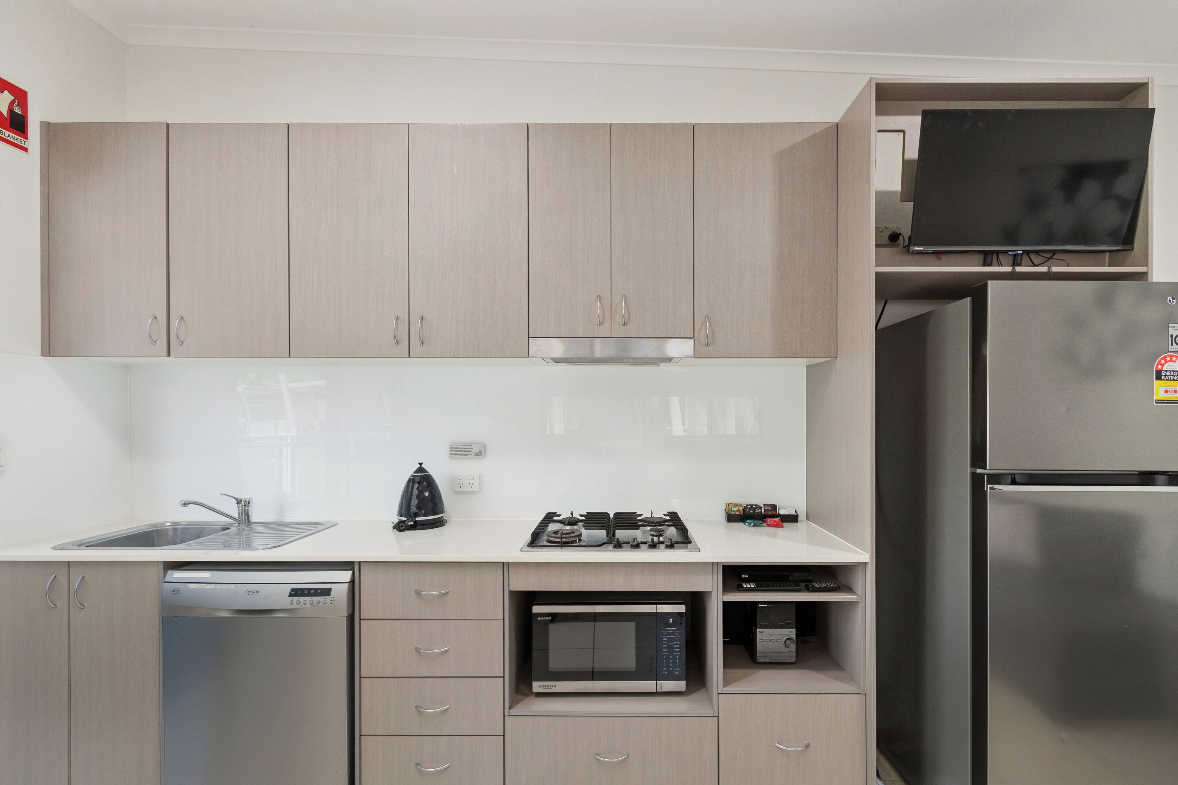 A modern kitchen with light wood cabinets, a stainless steel refrigerator, a built-in microwave, a dishwasher, and a flat-screen TV mounted above the stove.