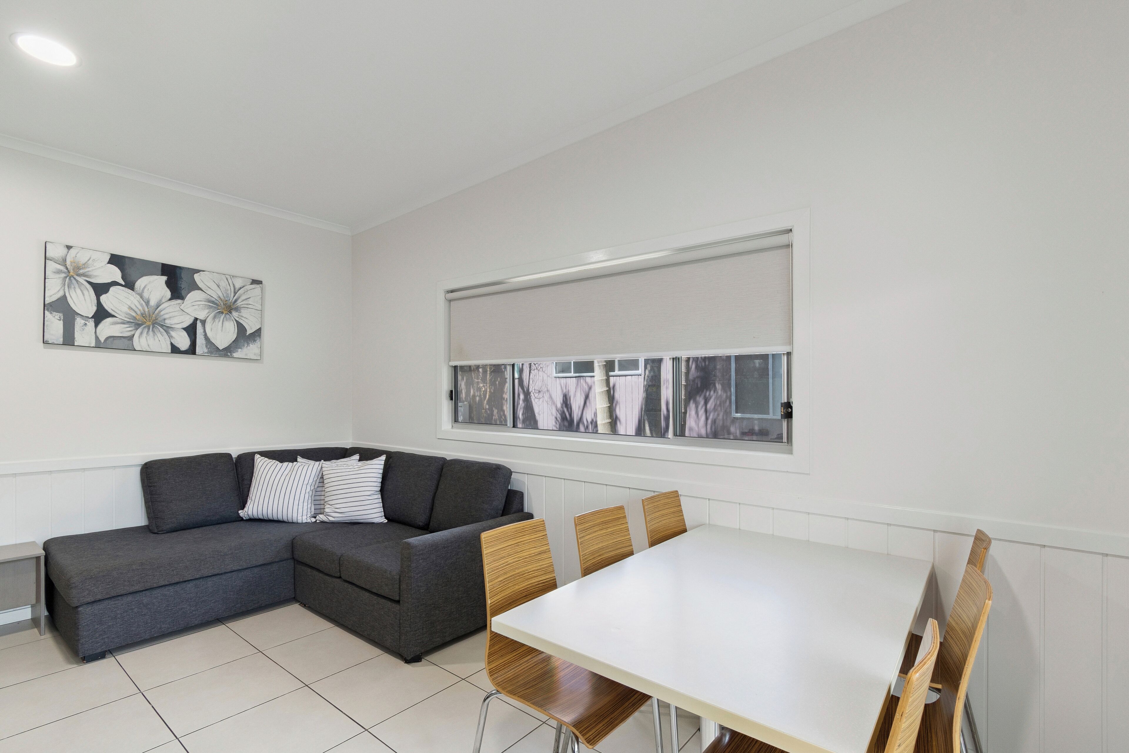 Un salon moderne avec un canapé d’angle gris, une table à manger blanche avec des chaises en bois, une grande fenêtre avec des stores et une œuvre d’art florale accrochée au mur.