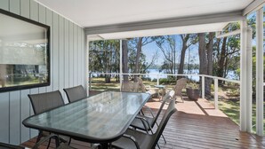 A covered outdoor deck with a glass table and chairs, overlooking a lake and surrounded by trees.
