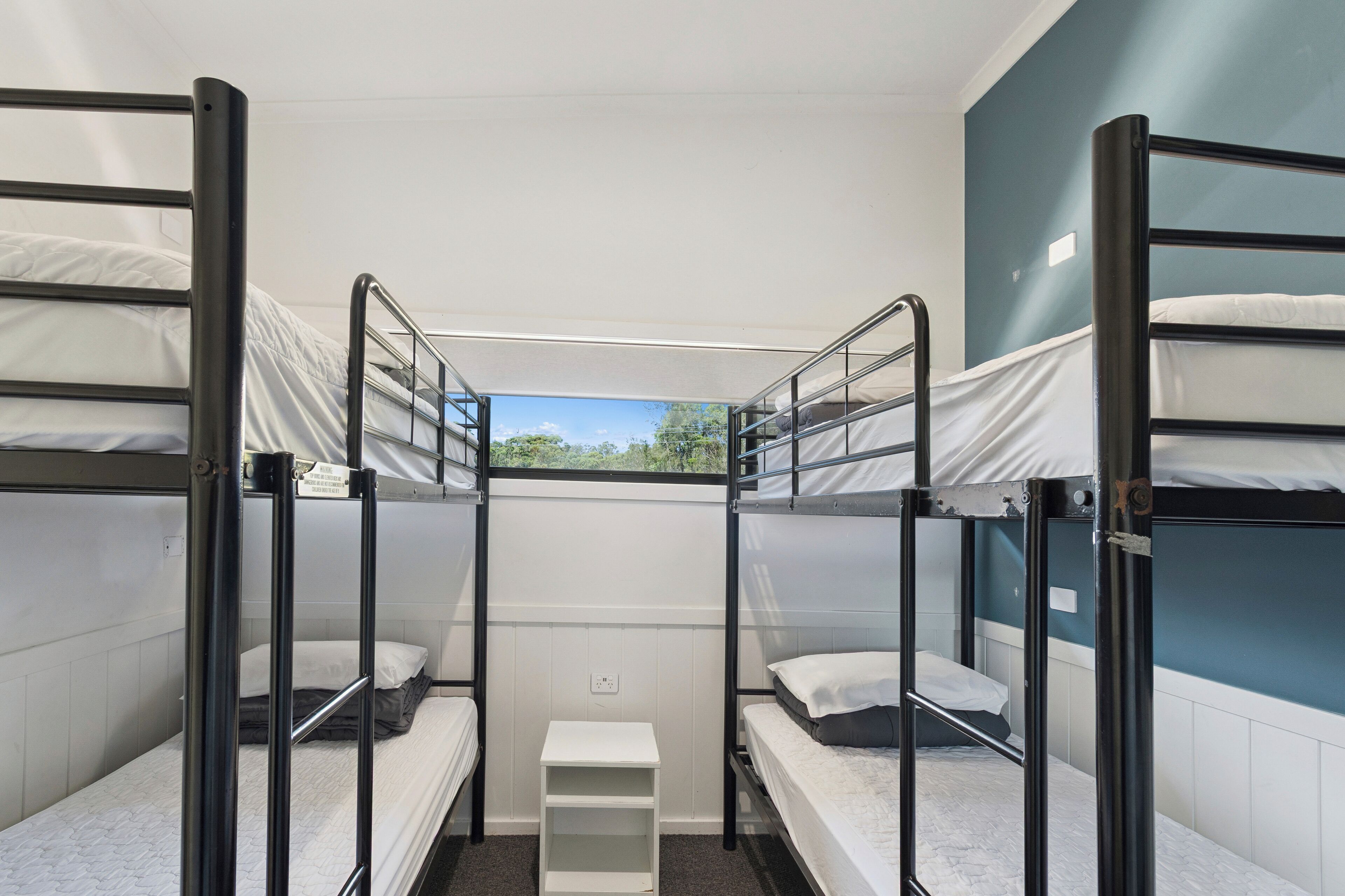 A bunk bed room with two beds, a small white nightstand, and a window with a view of trees.