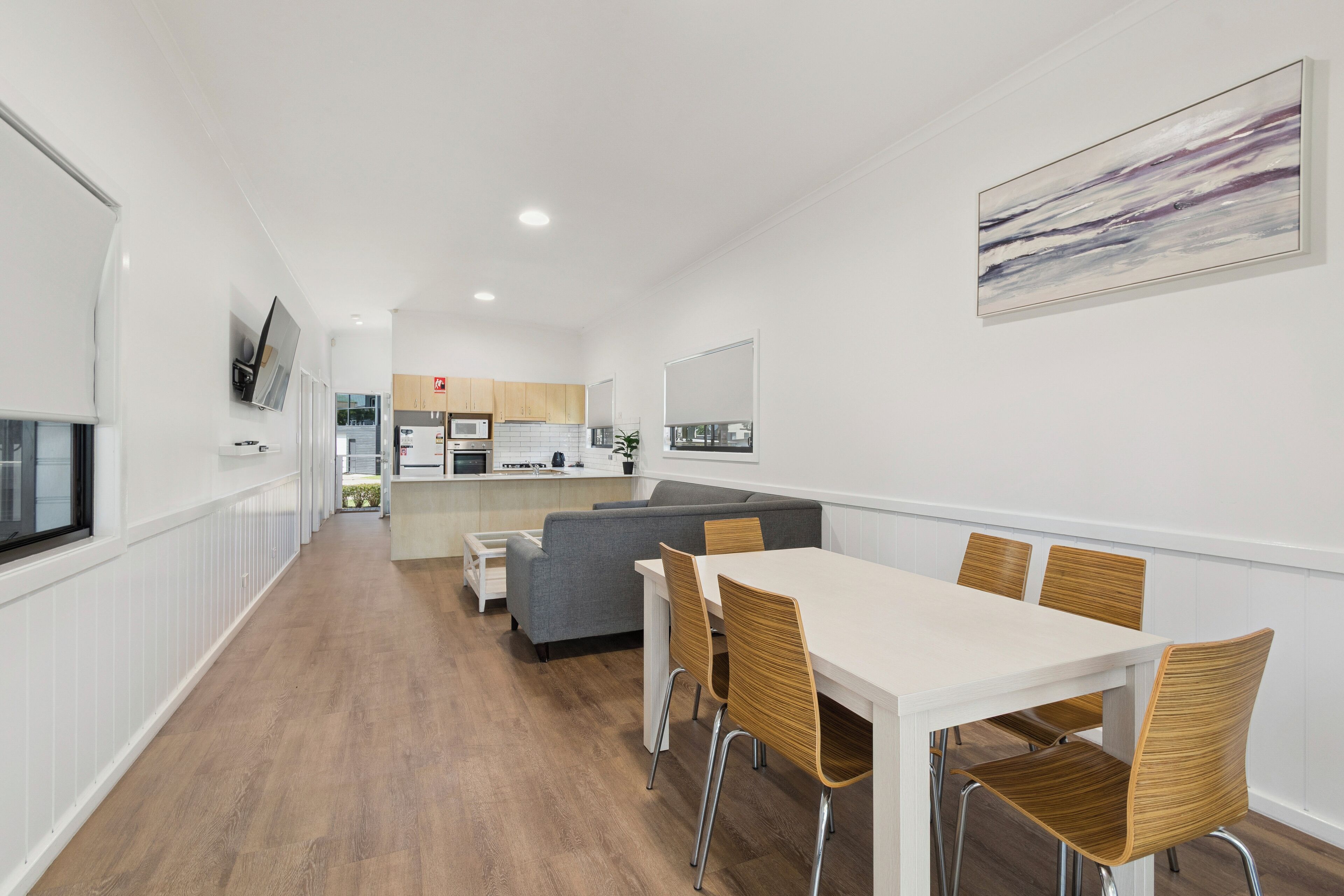 A modern kitchen with white cabinets, a grey sofa, and a dining area with a white table and wooden chairs.