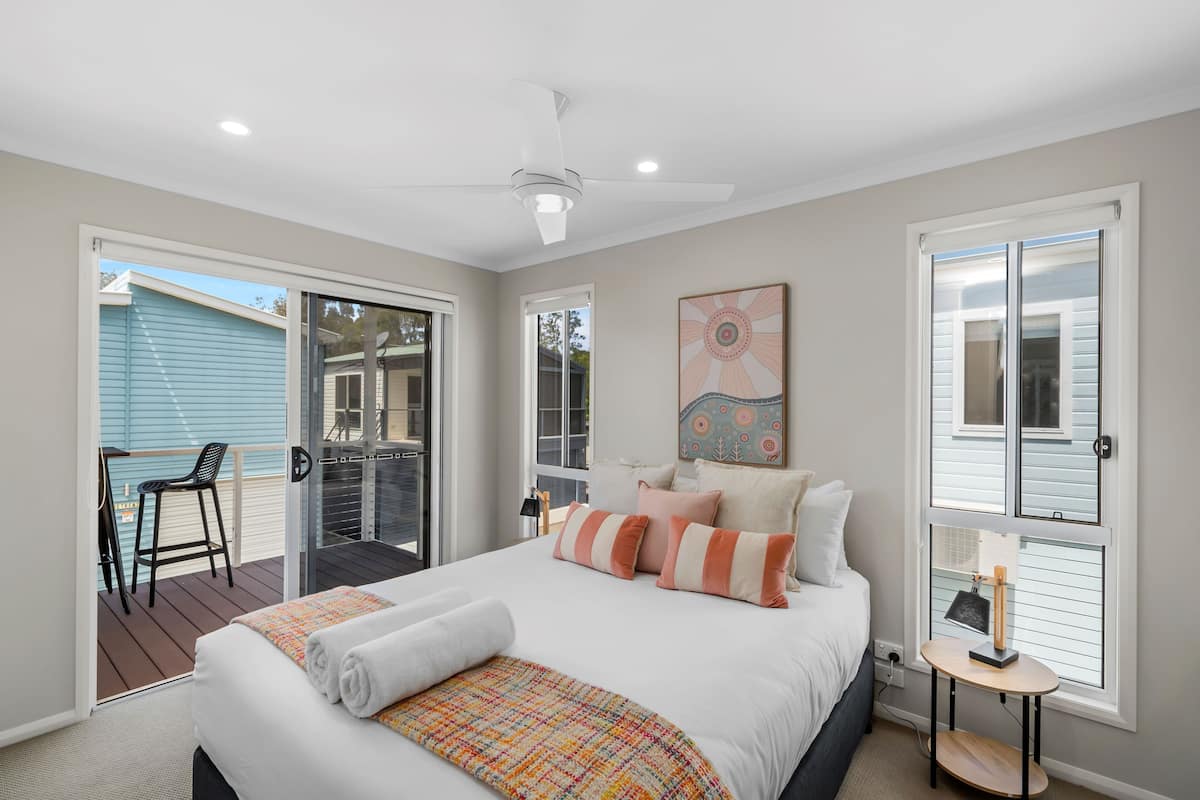 Une chambre avec un grand lit, un ventilateur de plafond, une fenêtre avec des stores et une vue sur une maison bleue à l’extérieur.