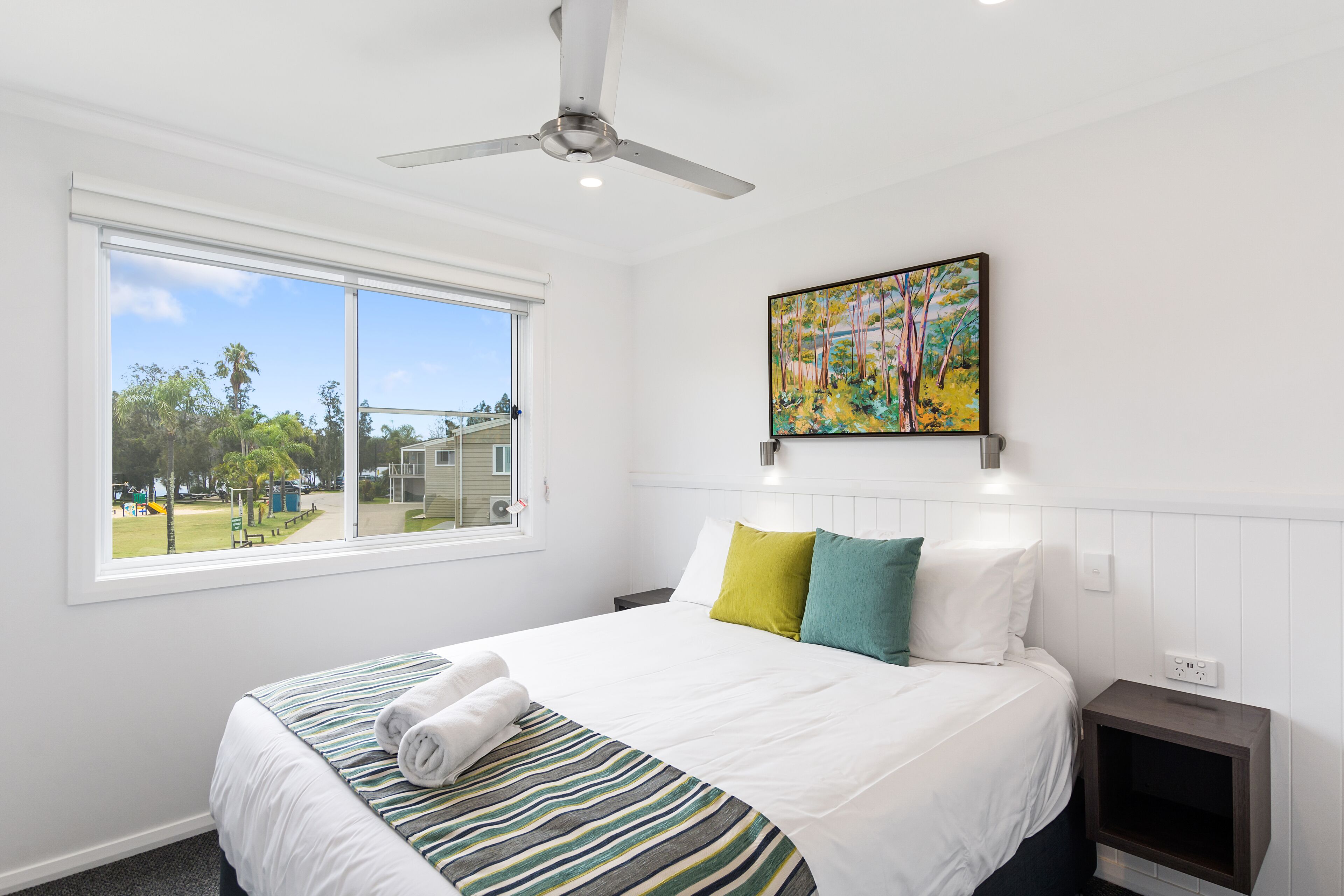 Habitación de hotel con una cama grande, ventilador de techo, ventana con vistas al exterior y un cuadro en la pared.
