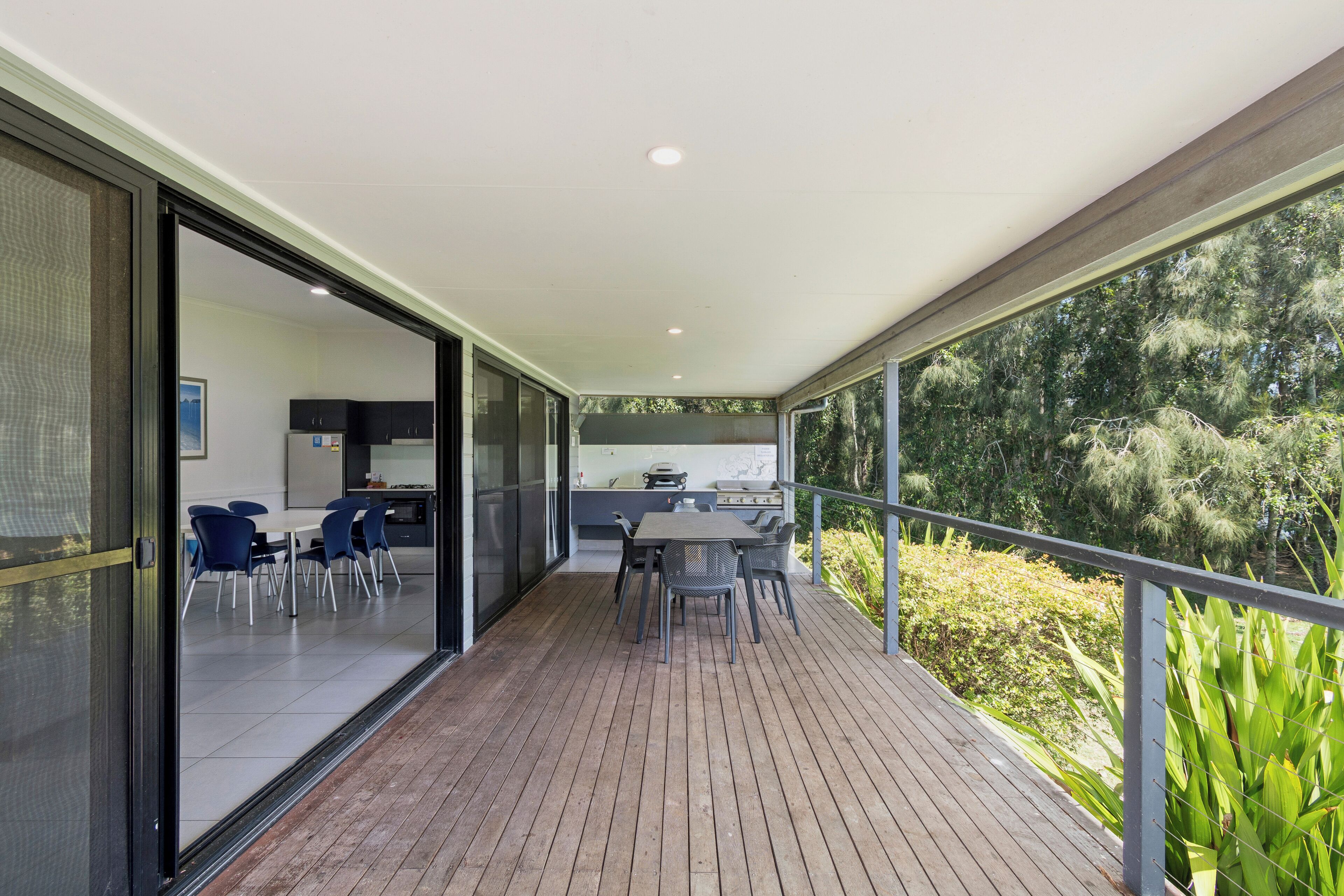 Un patio exterior moderno con terraza de madera, una mesa de comedor con sillas y una cocina visible a través de puertas de cristal.