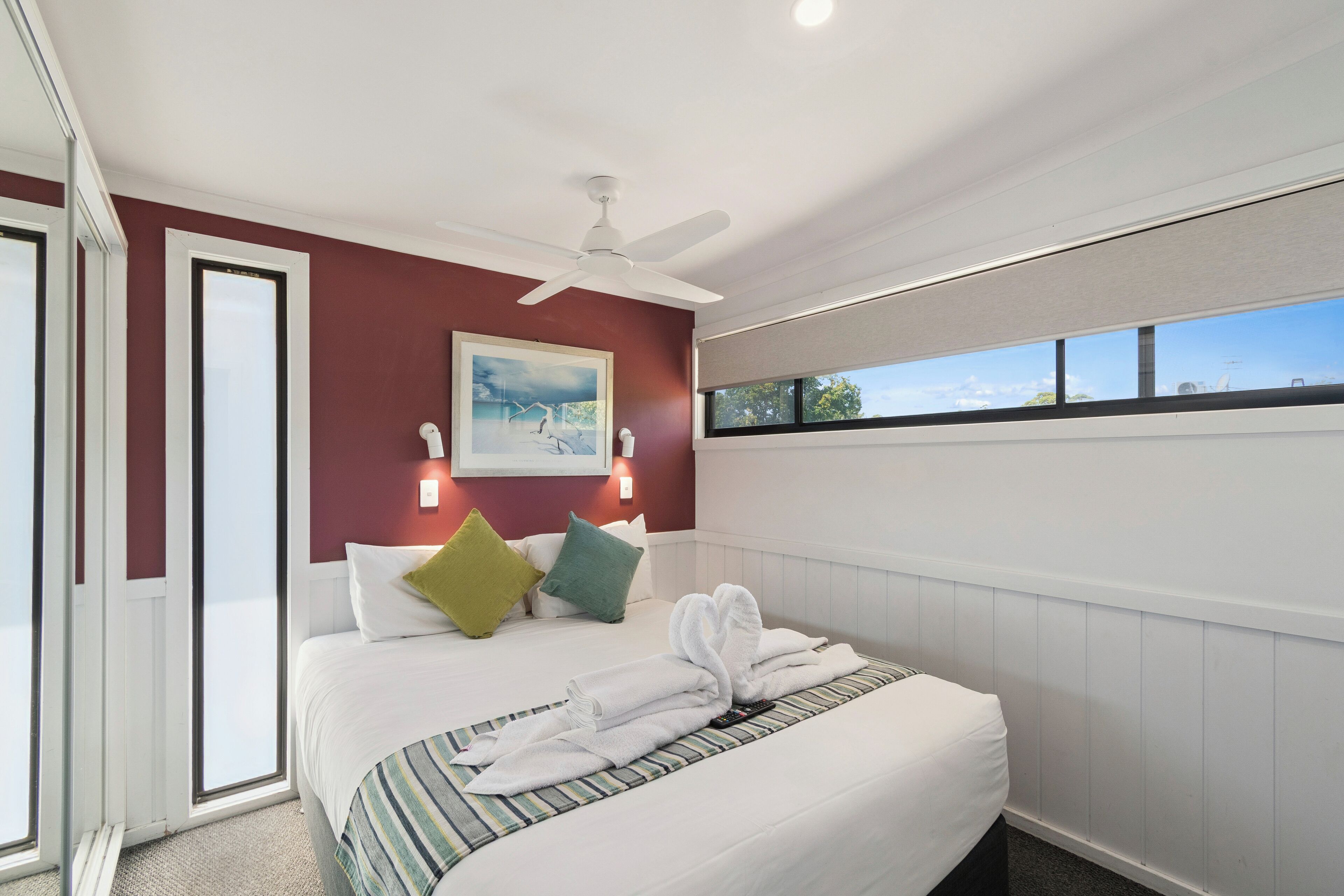 A hotel room with a bed, a ceiling fan, a painting, and a window with a view.