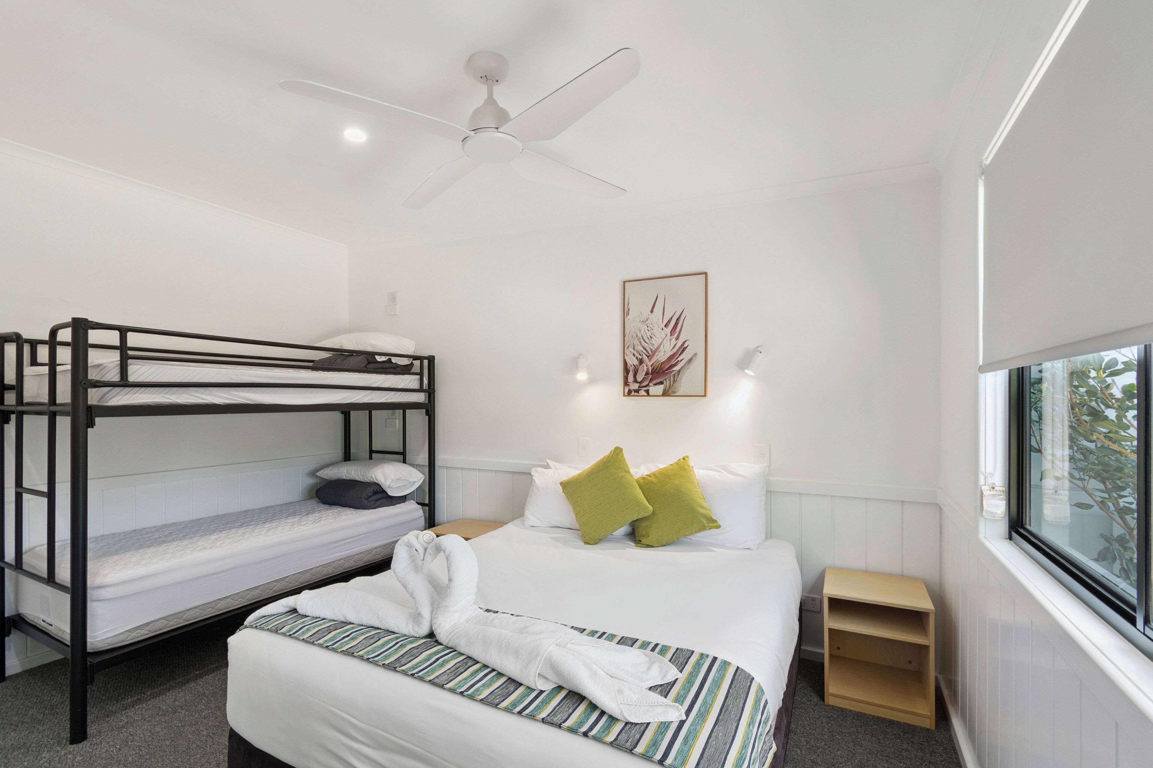 Quarto de hotel com cama de beliche, uma cama individual, ventoinha no teto, mesa de cabeceira, janela com vista e uma obra de arte emoldurada na parede.