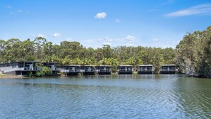 A row of modern, two-story buildings with balconies, situated by a calm lake, surrounded by dense greenery.
