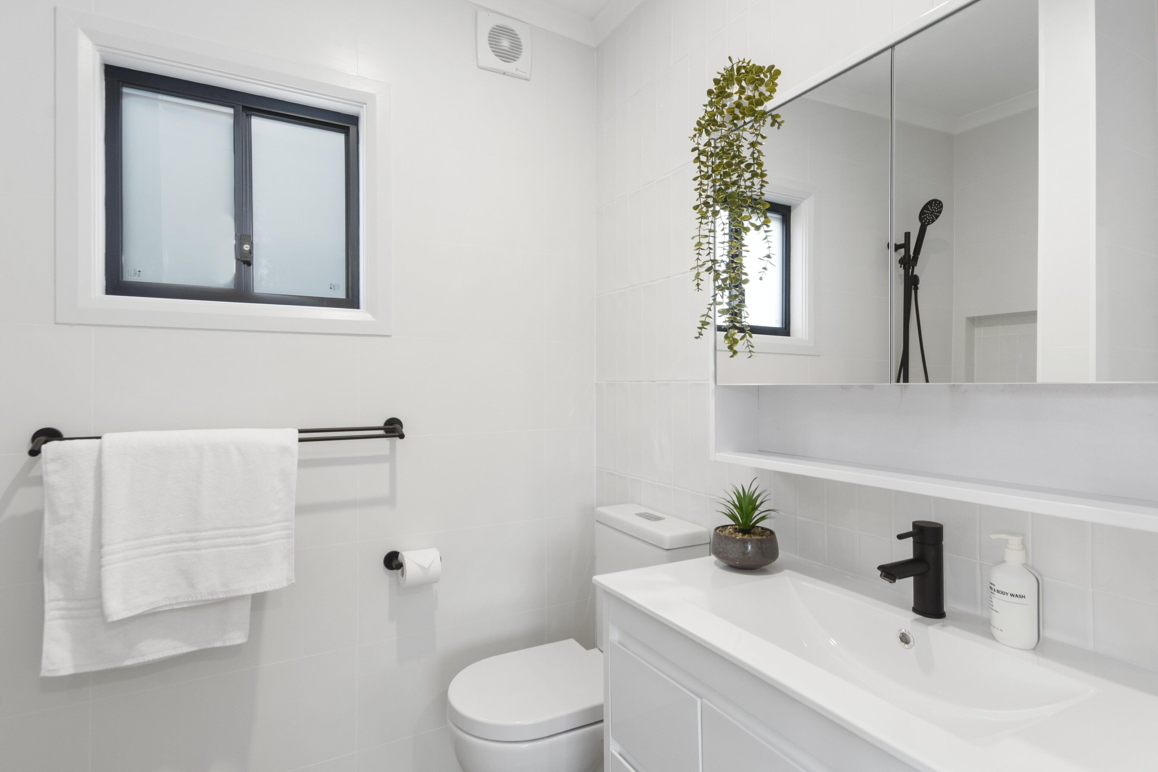 A modern bathroom with a white toilet, a sink with a mirror above it, a towel rack, and a window.