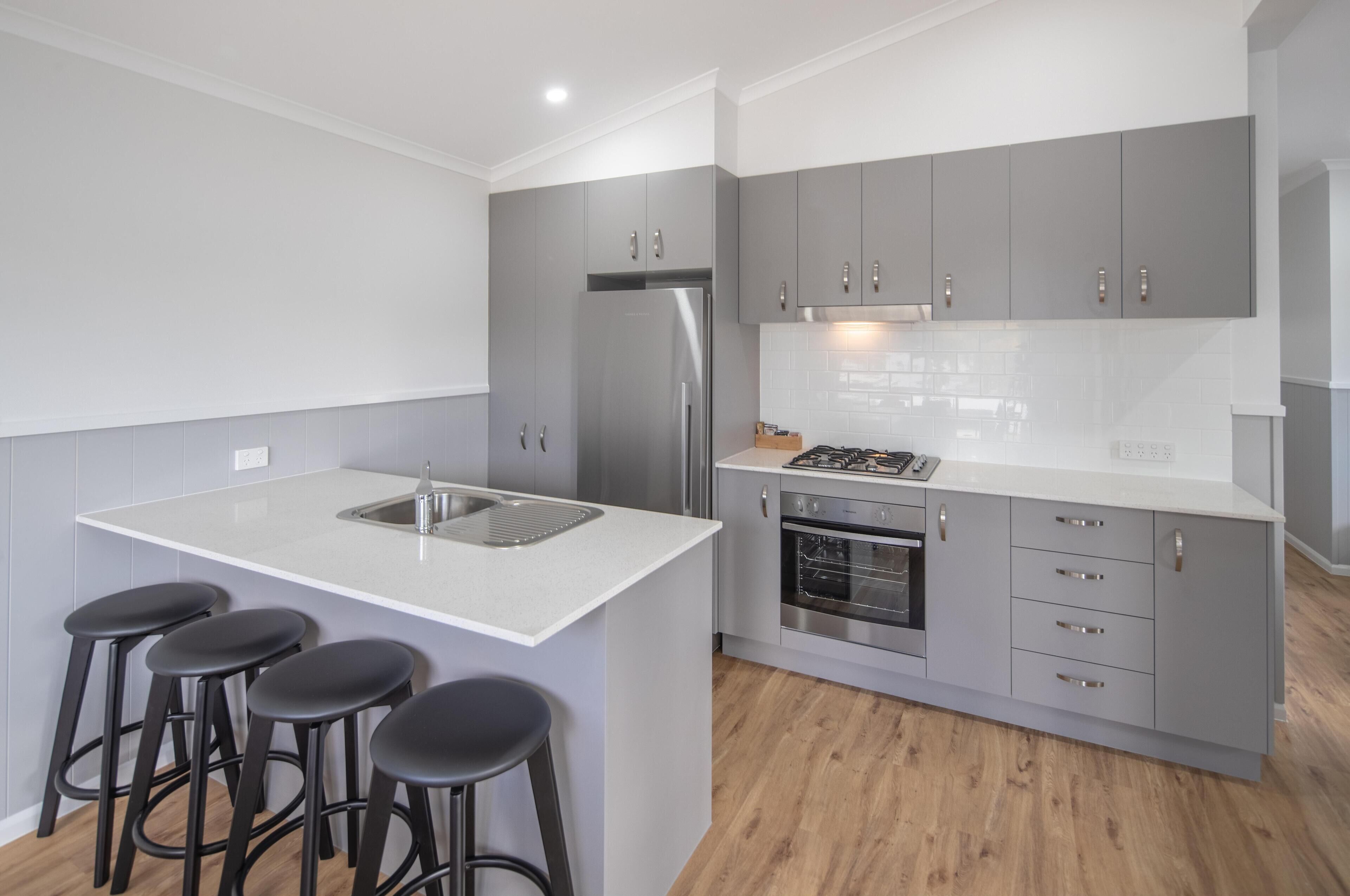 A modern kitchen with grey cabinets, a stainless steel refrigerator, a built-in oven, and a white countertop with bar stools.