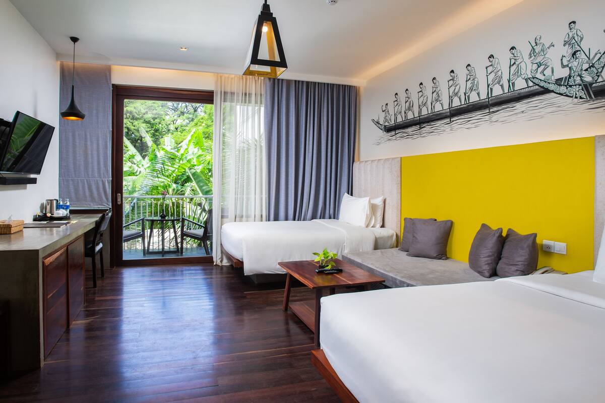 A hotel room with a bed, a sofa, a desk, and a TV. There is a balcony with curtains and a view of greenery.