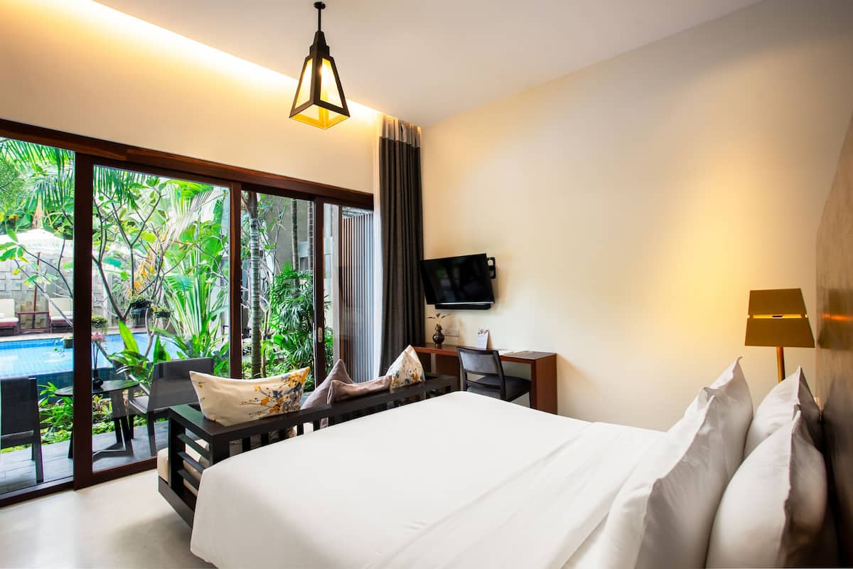 A hotel room with a bed, a desk, a chair, a TV, and a view of a swimming pool and greenery.