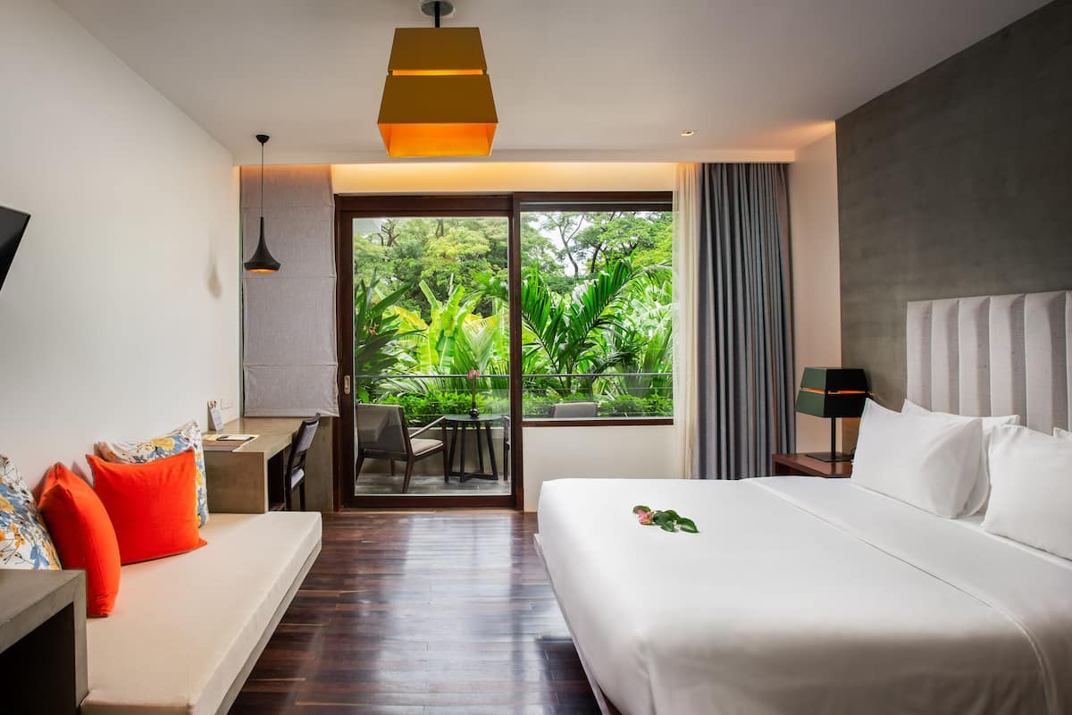 A hotel room with a bed, a TV, a sofa, a desk, and a balcony with a view of greenery.