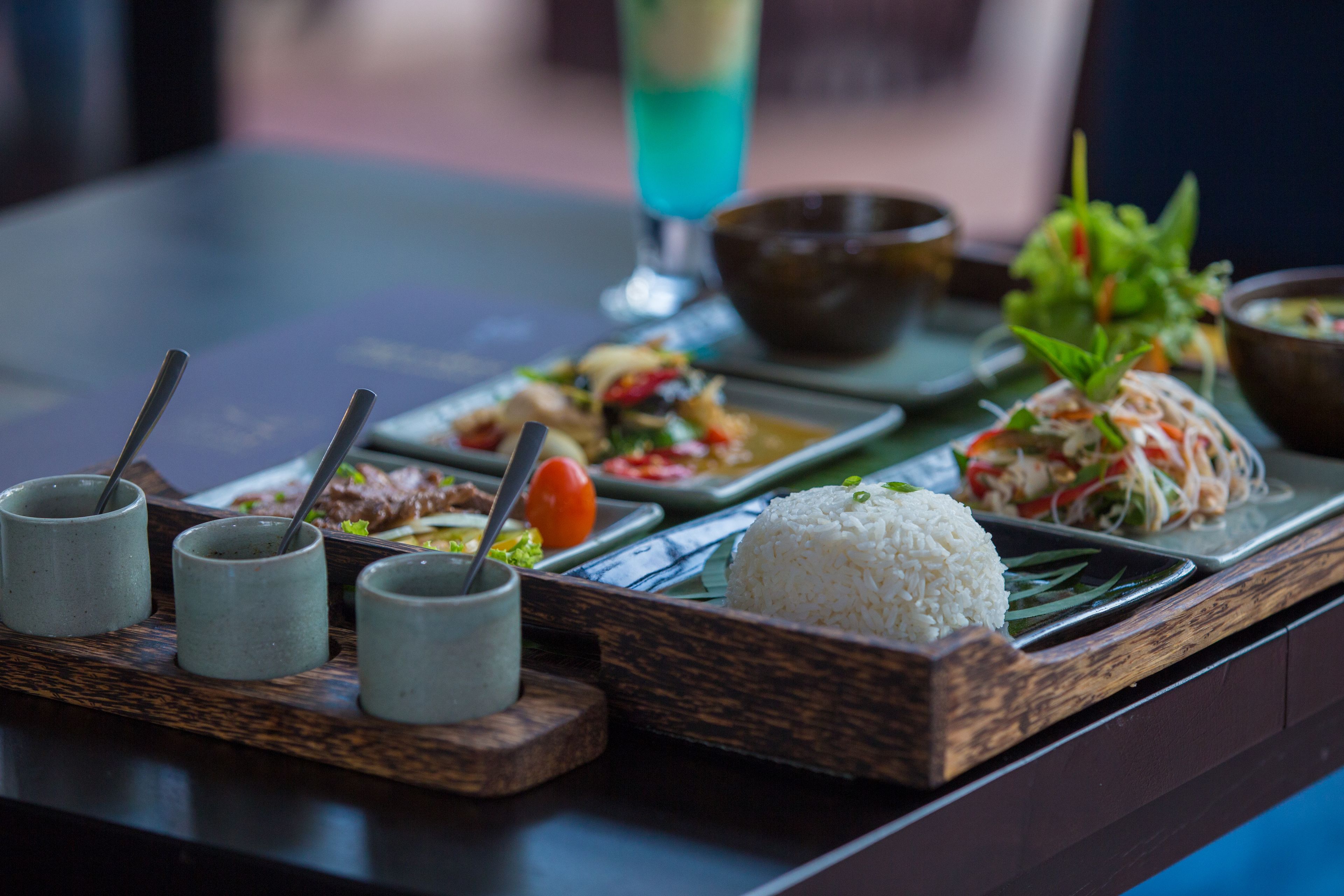 A tray with various dishes including rice, vegetables, and a blue drink on a table.