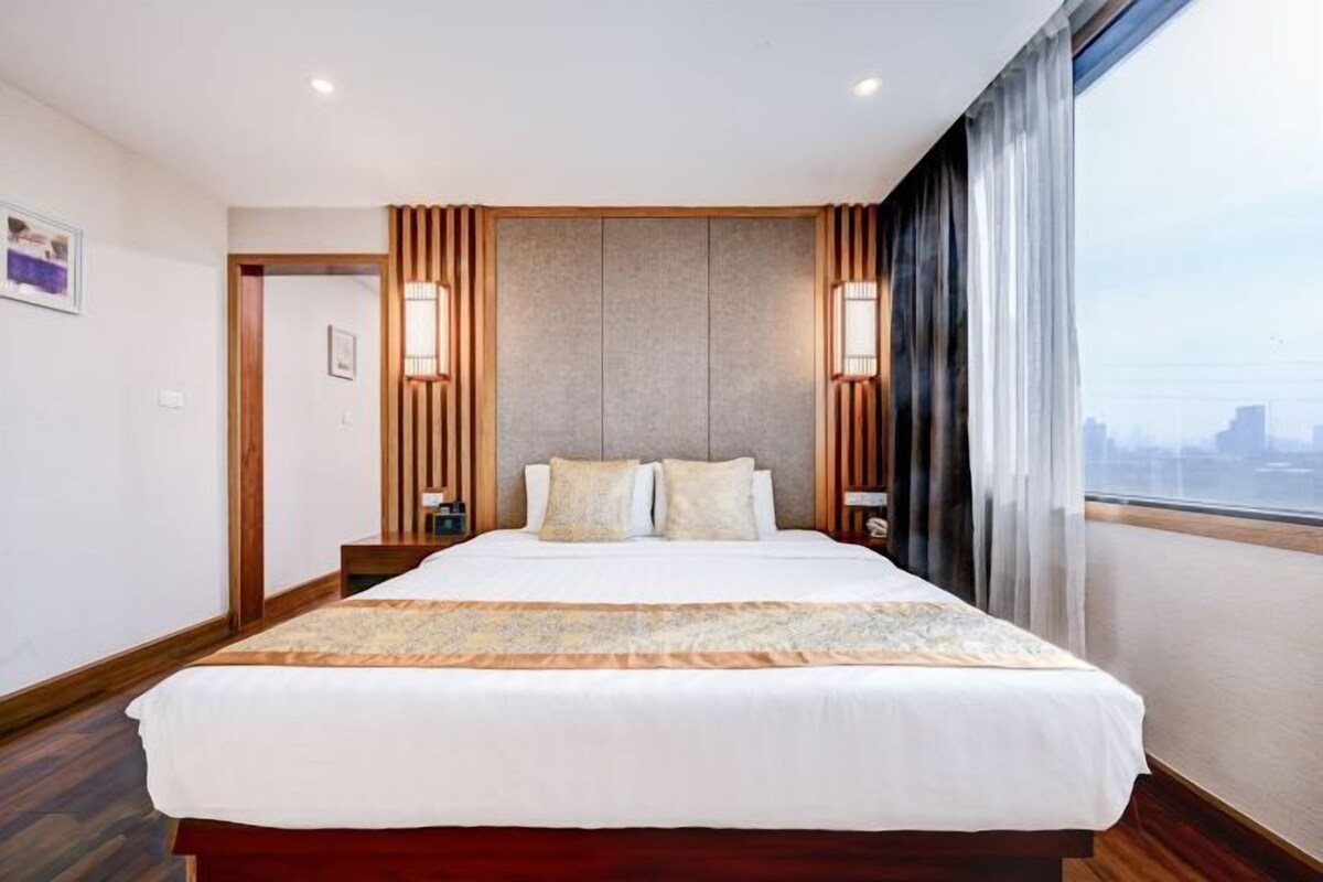 Una habitación de hotel con una cama grande, paneles de madera, vistas a la ciudad y una mesita de noche con una lámpara.