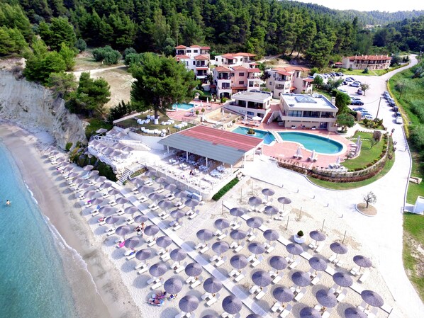 An aerial view of a resort with multiple buildings, a large pool, and a beach area with umbrellas and sun loungers.