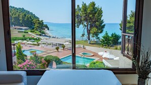 A hotel room with a large window overlooking a pool, beach, and forested hills. - Elani Bay Resort (Kassandra)