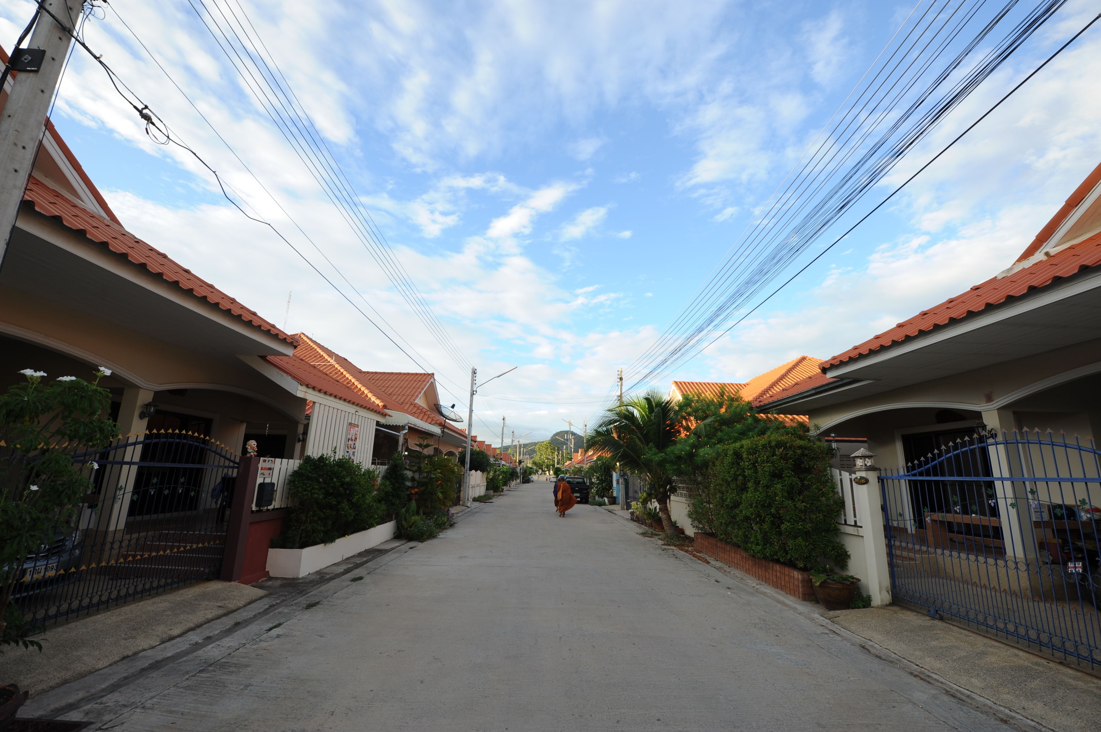 A residential street with houses on both sides, featuring red-tiled roofs and a clear sky.