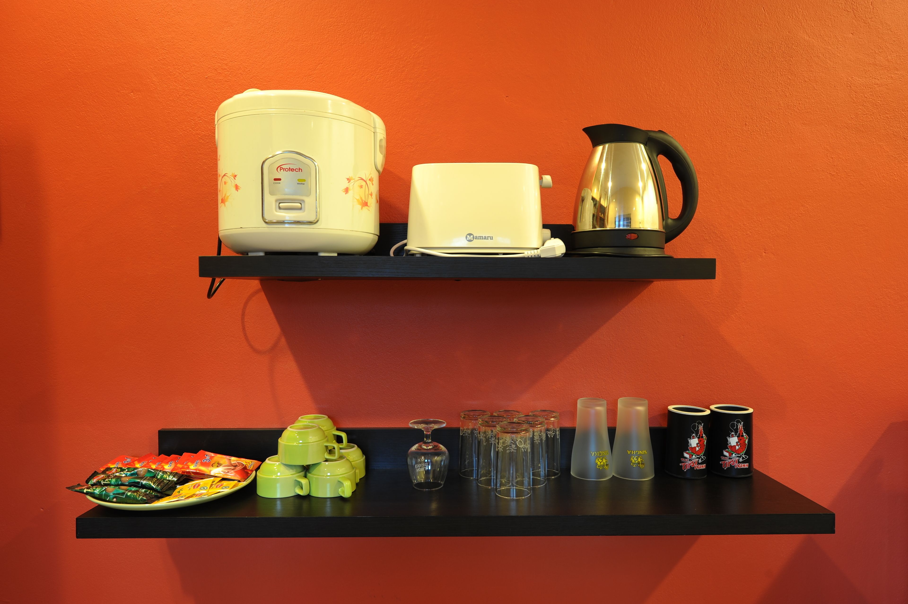A kitchen area with a rice cooker, toaster, kettle, and various kitchenware on shelves against an orange wall.