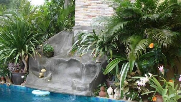 A swimming pool with a waterfall feature, surrounded by lush tropical plants. - Pius Place (Pattaya)