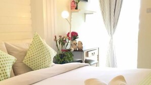 A neatly made bed with green polka dot pillows, a bedside table with a vase of flowers, and a view of a bright room through sheer curtains. - Pius Place (Pattaya)