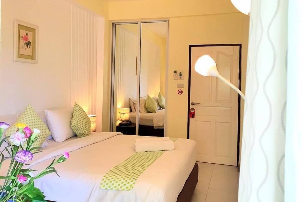 A hotel room with a bed, a mirror, a door, and a nightstand with a lamp.