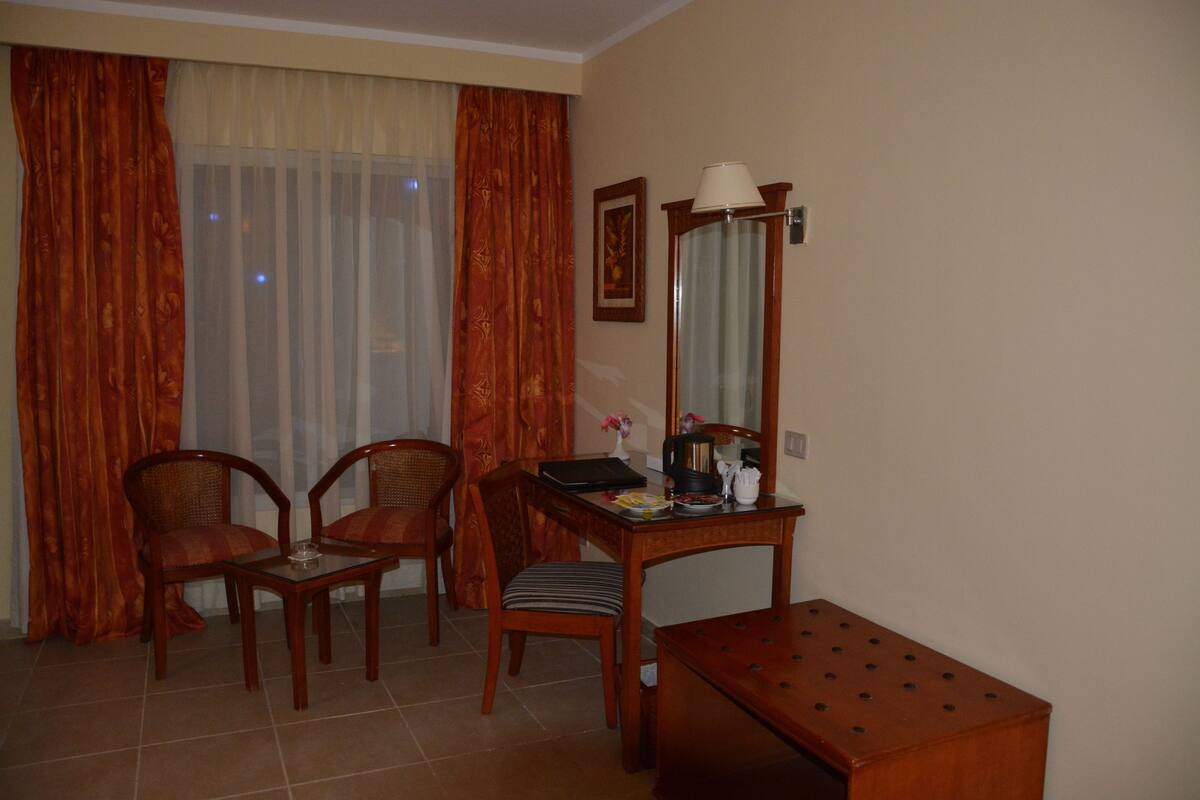 A hotel room with a wooden desk, two chairs, a small table, a mirror, and a window with curtains.