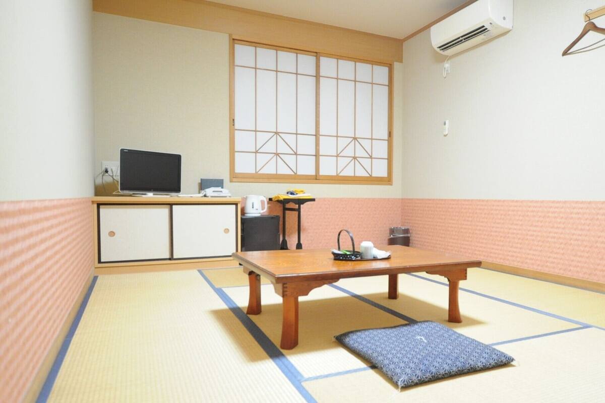 A room with tatami flooring, a wooden table, a television on a cabinet, and a window with a grid pattern.