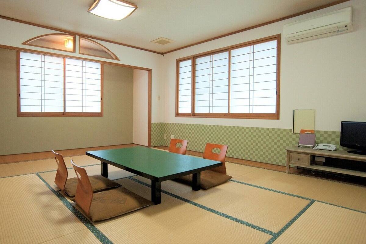 A room with tatami flooring, a low table with cushions, a television, and a small desk with a lamp.