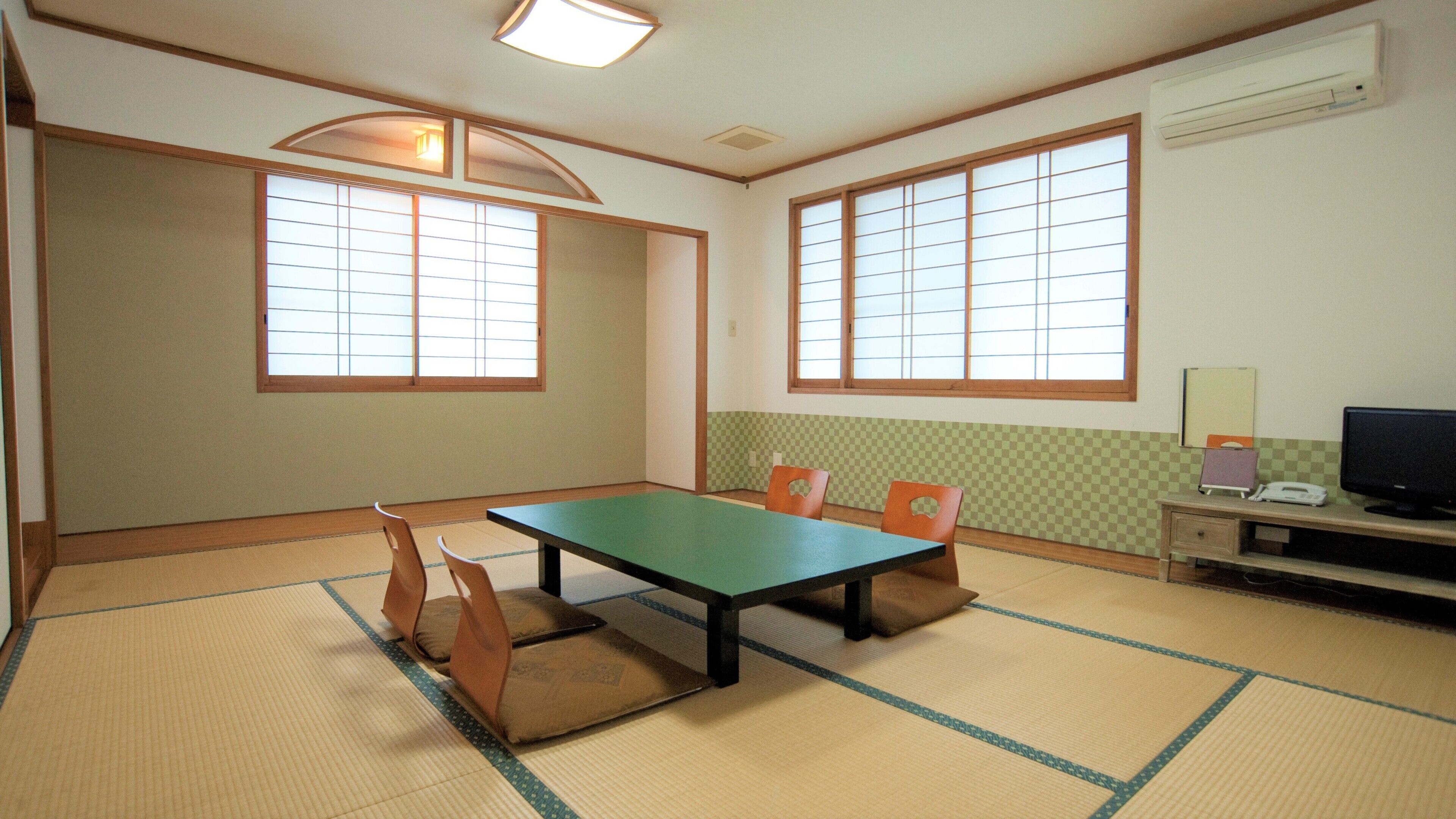 Una habitación con suelo de esteras de tatami, una mesa baja y sillas. Hay dos ventanas con marcos de madera y pantallas de papel. Un escritorio con una computadora y monitor está contra la pared.