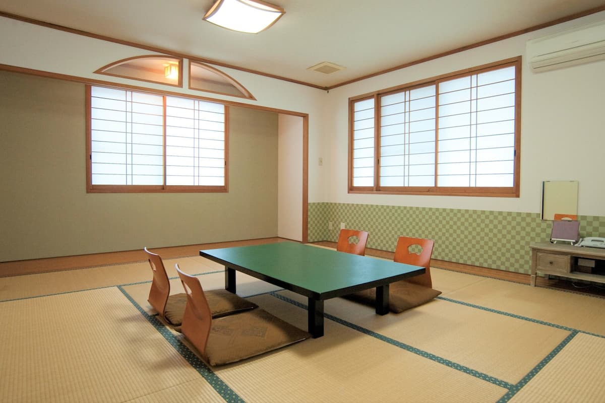 A room with tatami mat flooring, a low table, and chairs. There are two windows with wooden frames and paper screens. A desk with a computer and a monitor is against the wall.