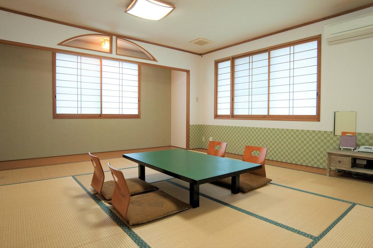 A room with tatami mat flooring, a low table, and chairs. There are two windows with wooden frames and paper screens. A desk with a computer and a monitor is against the wall.