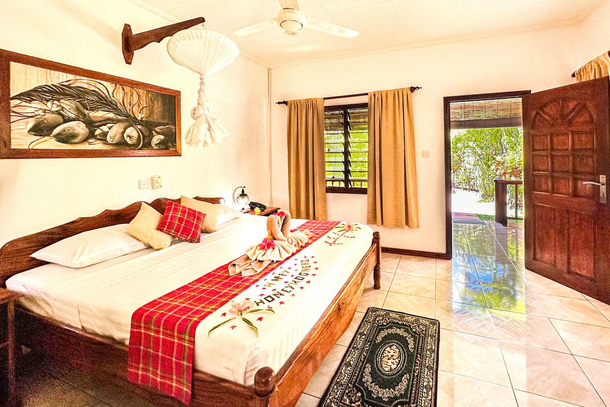 Un dormitorio con cama, un cuadro, ventilador de techo, ventana con cortinas y una puerta.