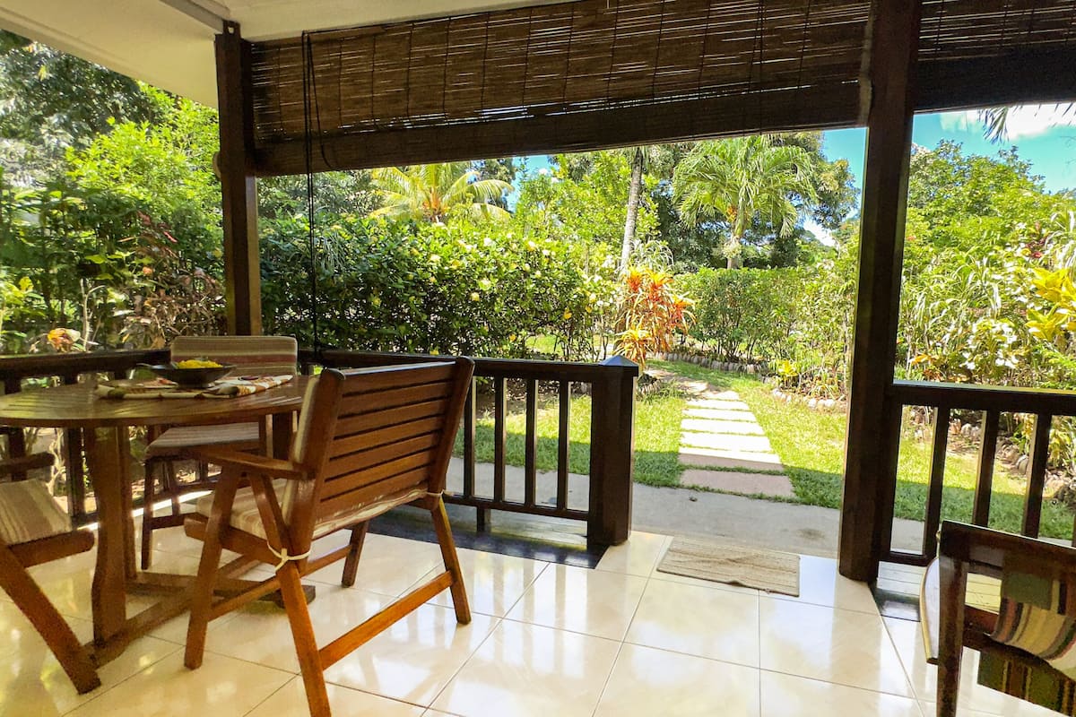 Un patio exterior con mesa y sillas de madera, rodeado de exuberante vegetación y un sendero que conduce a un jardín.
