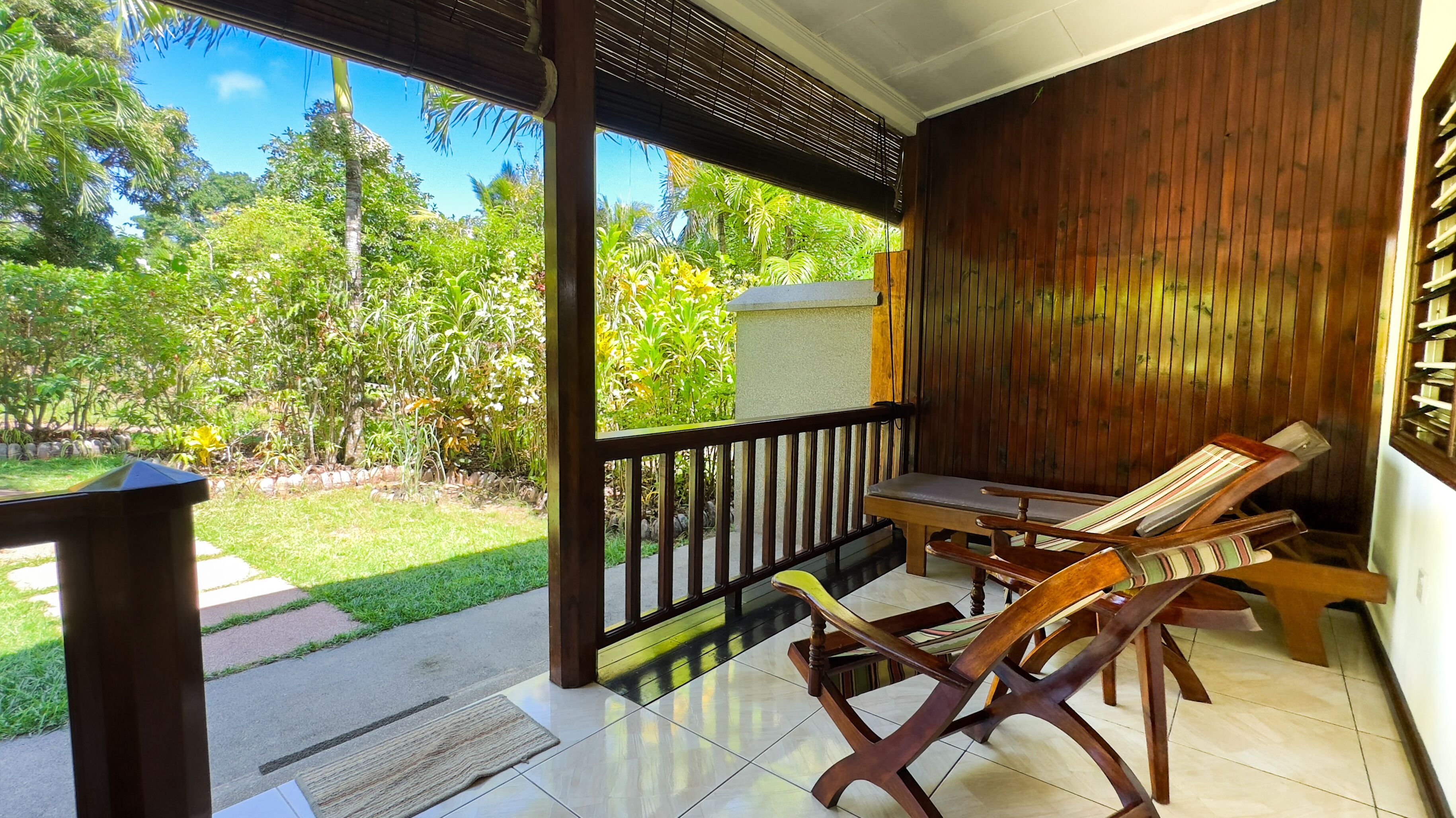 A covered patio area with a wooden table and chairs, a stone bench, and a view of a lush garden.
