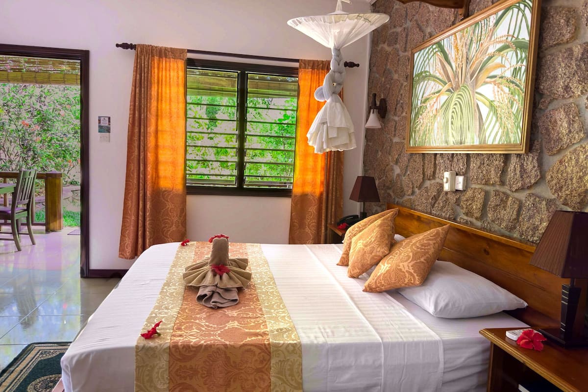 Una cama bien hecha con una toalla decorativa y una frutera, una pared de piedra con una planta enmarcada, y una ventana con cortinas.