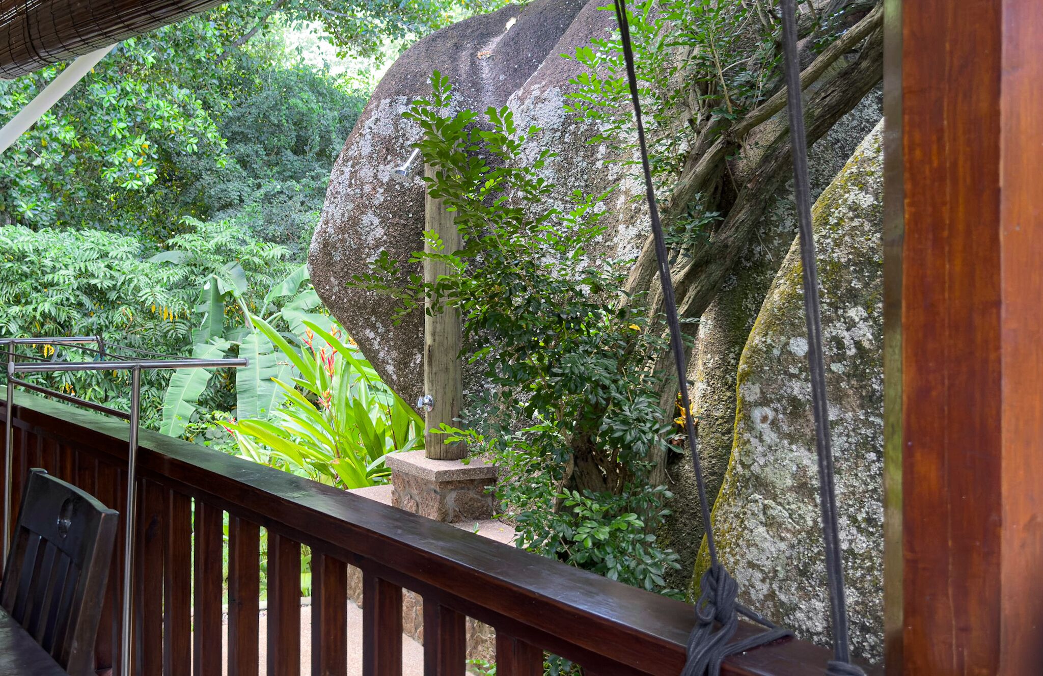 A wooden deck with a metal railing overlooking a lush garden with large rocks and dense foliage.