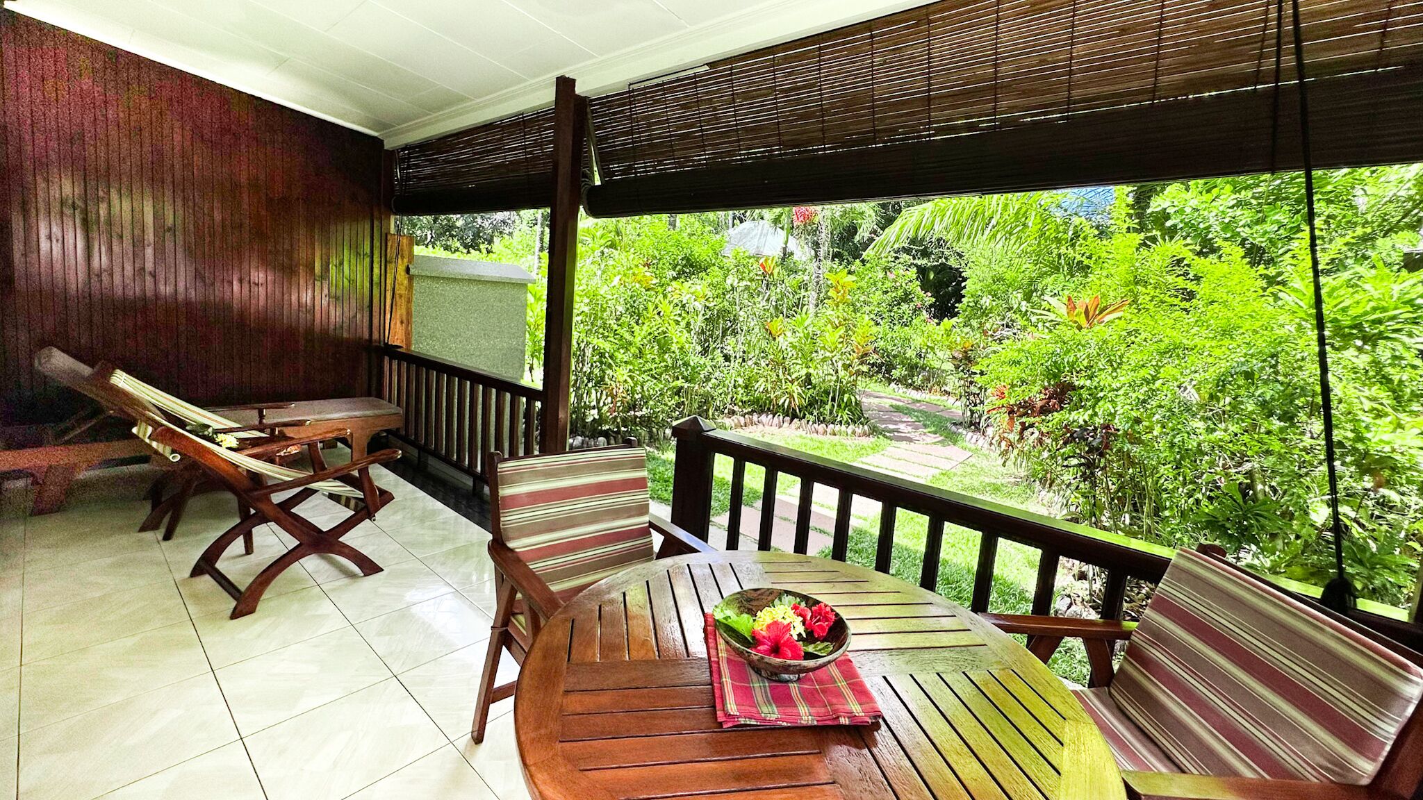 Um deck de madeira com mesa e cadeiras, uma tigela de frutas e vista para um verde exuberante.
