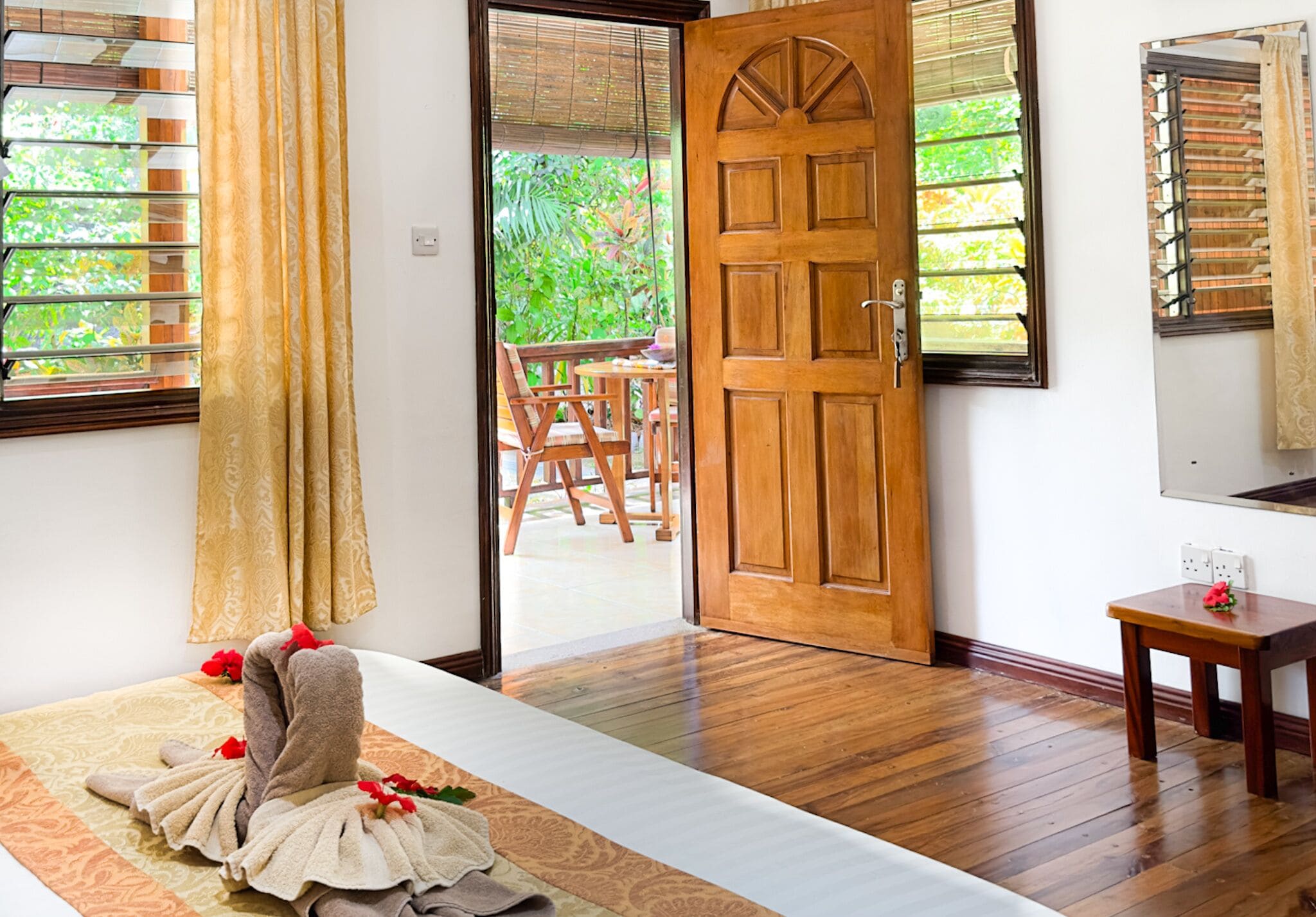 A hotel room with a wooden door, a bed with a patterned bedspread, a small wooden table with a flower, and a view of an outdoor area with a table and chairs.