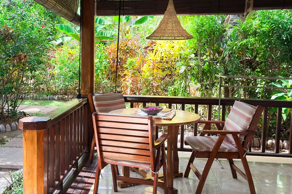 Un patio exterior con una mesa y sillas de madera, rodeado de abundante follaje y una lámpara colgante.