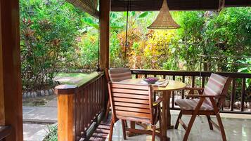 Un patio exterior con una mesa y sillas de madera, rodeado de abundante follaje y una lámpara colgante.