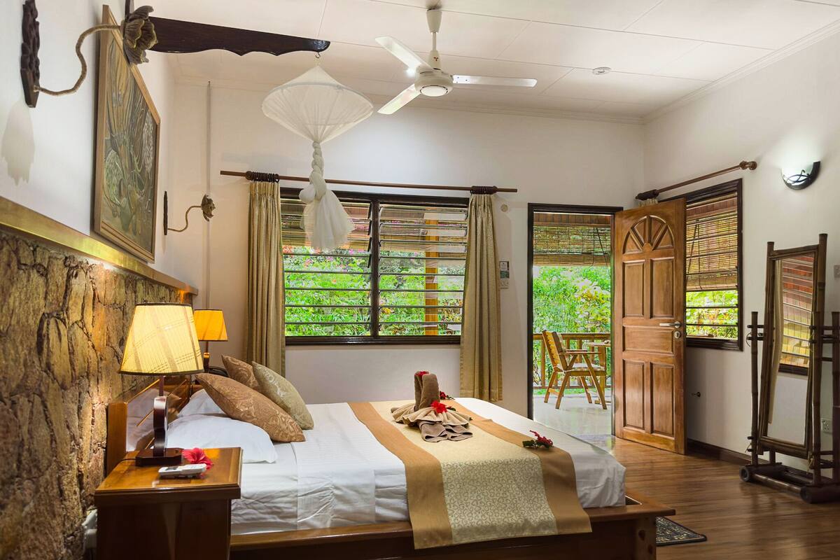 Un dormitorio con cama, mesita de noche, lámpara y vistas a la vegetación por la ventana.