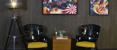 A room with two black chairs with yellow cushions, a wooden side table with a plant and books, and two abstract paintings on the wall.