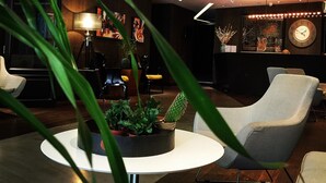 Een modern interieur met een ronde witte tafel, een cactus en een stoel. Er hangt een klok aan de muur en er staat een lamp op een statief.