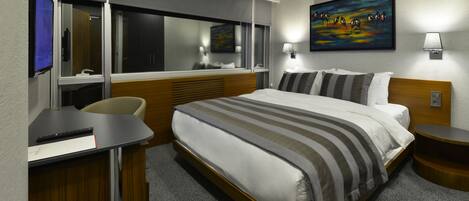A modern hotel room with a large bed, a desk, a chair, a TV, and a painting on the wall.