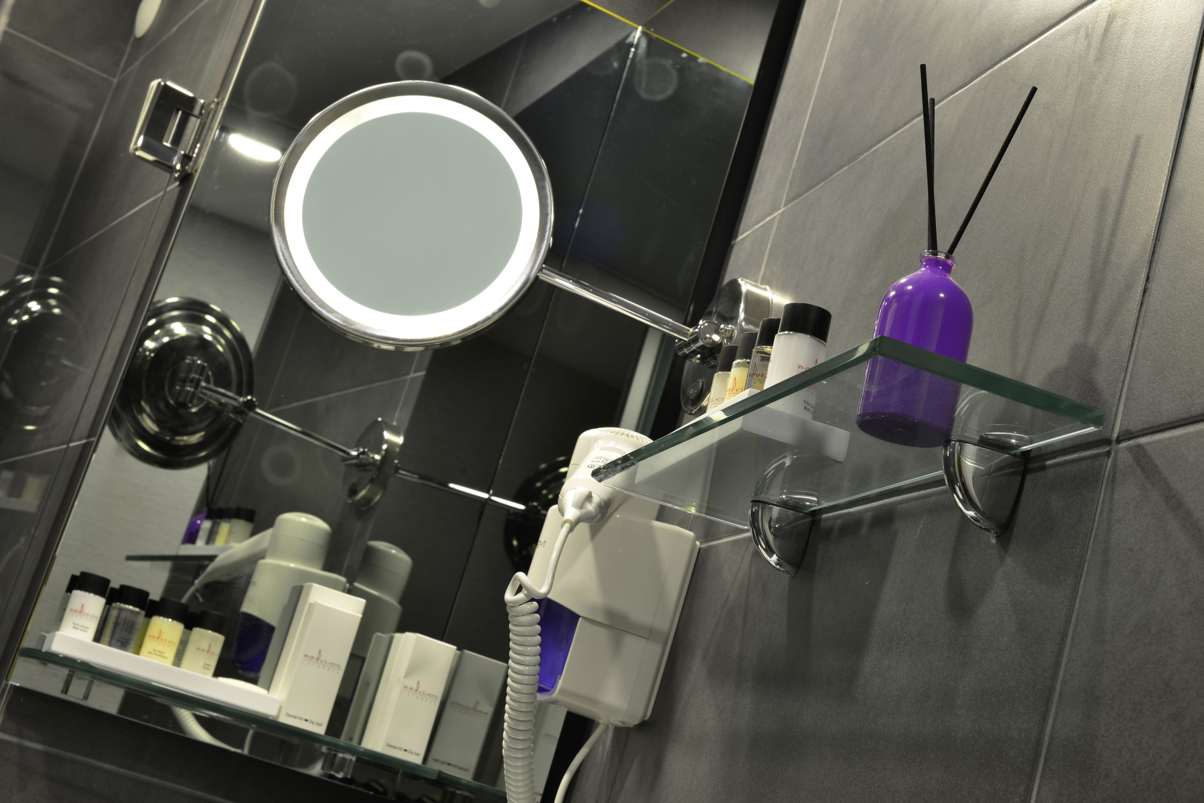 Ein modernes Badezimmer mit einer Glasablage für Pflegeprodukte, einem runden Spiegel und einer violetten Duftsprayflasche.